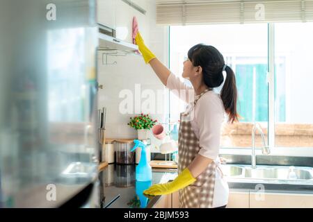 Donna asiatica che indossa guanti di protezione in gomma pulizia armadi da cucina nella sua casa durante il soggiorno a casa utilizzando il tempo libero circa la loro casa quotidiana Foto Stock