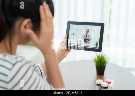 Vista posteriore della giovane donna che effettua una videochiamata con il suo medico con la sua sensazione di mal di testa sul tablet digitale online Healthcare tecnologia digitale servizio co Foto Stock