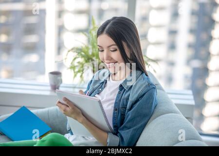 Giovane donna in una giacca di jeans seduta sul divano e prendere appunti Foto Stock