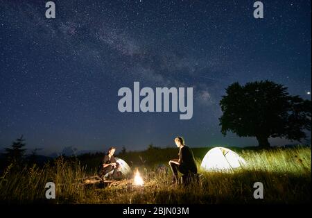 Escursionisti seduti di fronte a un altro su panchina di tronchi, guardando il fuoco insieme accanto al campo e tende di notte. Silhouette di un grande albero sullo sfondo, Via Lattea e cielo stellato sopra di loro Foto Stock