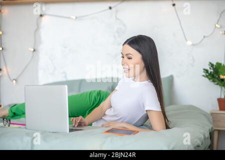 Giovane donna dai capelli scuri che si posa sul letto con un laptop Foto Stock