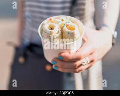 Gelato arrotolato in tazza di cono nelle mani della donna. Donna in abito a righe tiene una tazza conica con gelato a banana di kiwi in stile thailandese. Foto Stock
