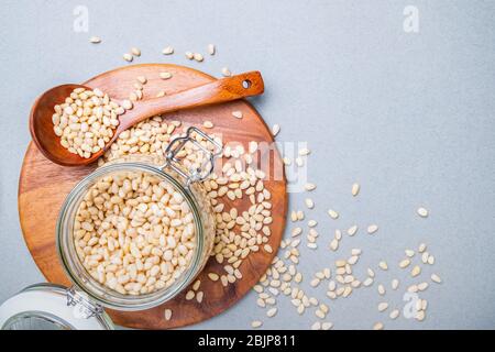 Layout. Vaso in vetro con noci di cedro siberiano su un piedistallo in legno su sfondo grigio. Nelle vicinanze si trova un cucchiaio di legno. Dadi sparsi sulla surfa Foto Stock