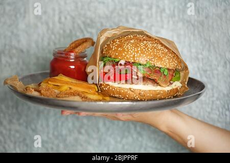 Donna con vassoio metallico di gustoso hamburger con prosciutto su sfondo grigio Foto Stock