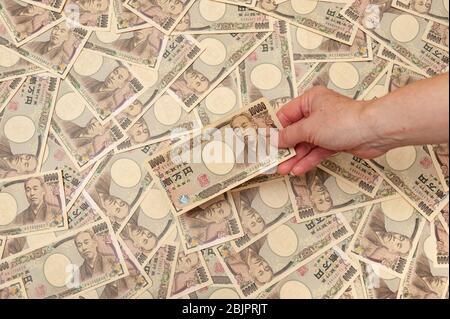 Mano che tiene una banconota da 10,000 yen - sfondo con diverse banconote da 10,000 yen (fronte). Denaro giapponese. Concetto: Abbondanza finanziaria. Foto Stock