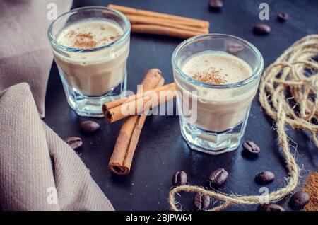 Liquore espresso su un tavolo. Liquore irlandese di crema su fondo scuro con chicchi di caffè e cannella. Bicchiere di liquore cremoso con caffè BE Foto Stock