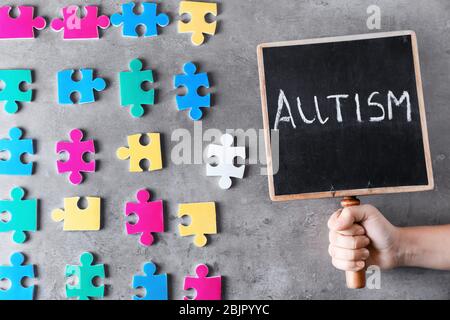 Donna che tiene lavagna con parola 'autismo' vicino pezzi colorati di puzzle su sfondo grigio Foto Stock