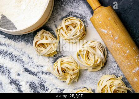 Varietà di spaghetti e tagliatelle di pasta cruda, fatti in casa, con farina su tavola di legno a dondolo per la pasta su pasta nera Foto Stock