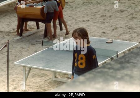 Un ragazzo di 11 anni in pantaloncini denim e una felpa blu scuro con un 18 giallo su di esso, camminando accanto ad un tavolo da ping pong sulla spiaggia a St Cast le Guildo, Bretagna, Francia 1974. Foto Stock