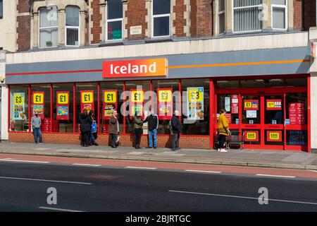 Gli acquirenti che effettuano le distanze sociali mentre fanno la coda fuori di un supermercato Islanda durante la pandemia del covid-19 Foto Stock