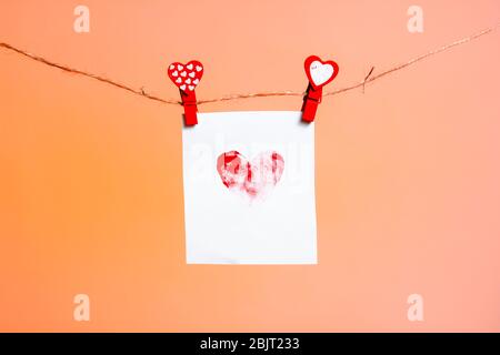 un pezzo di carta con un cuore modellato di impronte digitali di clothespins rossi con un cuore su una corda. San Valentino amore. Lava sfondo lussureggiante. Copia sp Foto Stock