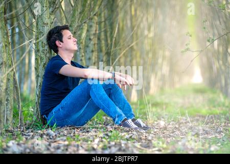 giovane seduto da un albero nel bosco che pensa alla vita e al riposo. spazio copia Foto Stock