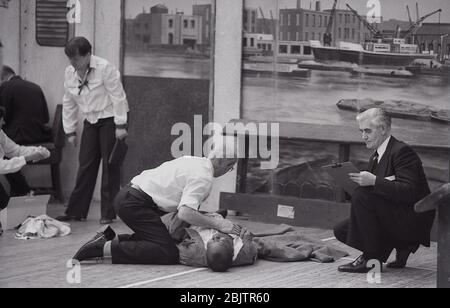 Anni '80, storico, all'interno di una sala, le persone di un corso di primo soccorso è segnato da esaminatori di funzionari sugli elementi pratici, con un primo soccorritore maschile nella foto che dà compressioni toraciche o CPR a un uomo che giace sul pavimento, Inghilterra, Regno Unito. Foto Stock