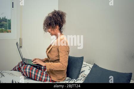 Vista laterale di una donna nera che lavora a casa durante la quarantena di Coronavirus. Chiamata in conferenza in camera da letto con pigiama e giacca. Foto Stock