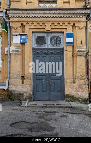 Muro di mattoni gialli di un edificio storico nella città di Kyiv. Vecchia porta in legno dipinta di grigio con finestre incorniciate ornate sopra. Esterno di un edificio retrò. A danneggiato Foto Stock