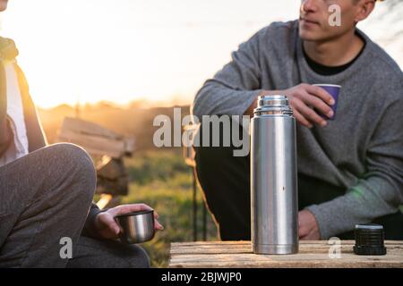 Indossare abiti casual uomo a bere tè caldo da un metallo thermos, mentre trascorrere pausa pranzo con la ragazza, luci del sole sullo sfondo Foto Stock