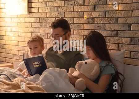 Padre che legge la Bibbia ai suoi bambini piccoli a letto Foto Stock