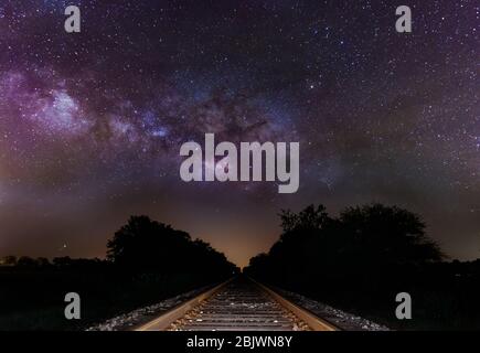 Milky Way si anima sulle piste del treno. Nucleo galattico che sorge maestosamente su binari ferroviari abbandonati in un campo rurale. Foto Stock