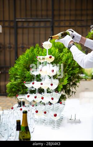 Una piramide di bicchieri con ciliegie da cocktail. Le mani di un cameriere con guanti bianchi versano champagne da una bottiglia. Una cascata di champagne e fumo bianco dentro Foto Stock