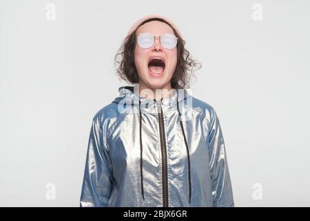 Donna emozionale arrabbiata con cappotto d'argento e occhiali da sole che urla. Emotivo, giovane faccia. Emozioni umane, concetto di espressione facciale. Foto Stock