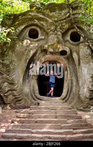 La turista femminile fa un volto spaventoso in piedi nella foce dell'Inferno al Parco dei Mostri, che fu commissionato nel 1552 dal Principe Pier Fr Foto Stock