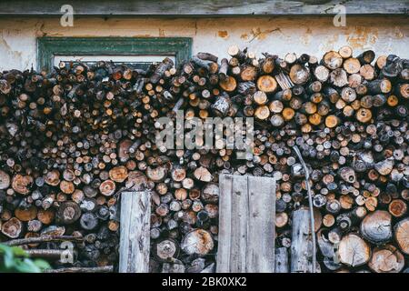 Pali di legno con legna da ardere sullo sfondo di un muro di casa chiude una finestra verde Foto Stock