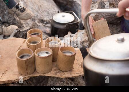 Versare il caffè caldo in tazze di bambù da un vecchio bollitore in metallo, in un ambiente roccioso sulla cima della montagna. Foto Stock