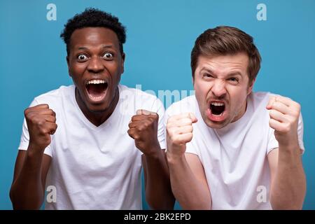 Felice due amici maschi in bianco t-shirt posa isolato su sfondo blu. Concetto di stile di vita sportivo e ricreativo. Coraggio di sostenere la squadra preferita, clenching Foto Stock