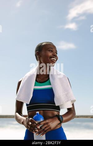 Donna sorridente con asciugamano intorno al collo e tenendo una bottiglia d'acqua che guarda via. Donna sportiva rilassante dopo una corsa all'aperto. Foto Stock