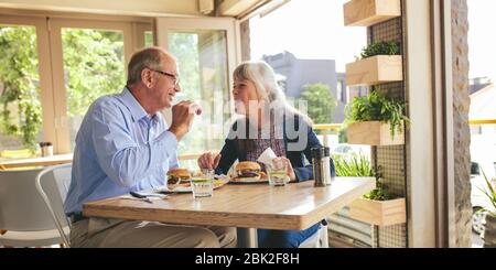Coppia senior che si divertendete a mangiare hamburger insieme in un bar. Sorridente uomo maturo e donna che mangiano hamburger in un ristorante. Foto Stock