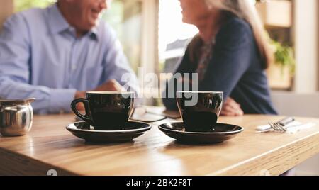Due tazze di caffè al tavolo con una coppia senior seduta sullo sfondo in un bar. Tazza di caffè fresco sul tavolo del caffè con uomo e donna in pensione seduti a. Foto Stock