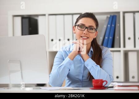 Donna d'affari in occhiali si siede in ufficio al tavolo mani tenendo sotto il mento e sorridendo Foto Stock