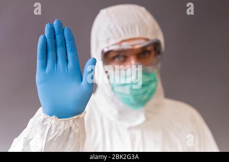 Medico maschile con maschera chirurgica, occhiali e tuta protettiva che mostra stop su sfondo grigio. Foto Stock