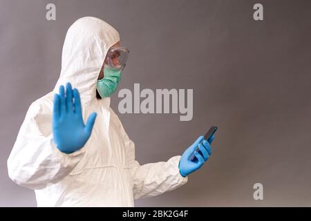 Medico con maschera chirurgica, occhiali e tuta protettiva che tiene il telefono e mostra stop con una mano su sfondo grigio. Foto Stock