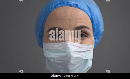 Medico femminile in maschera protettiva e cappuccio blu, faccia in primo piano isolata sulla parte posteriore grigia Foto Stock