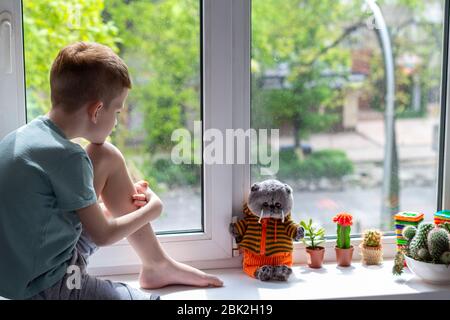 Ragazzo seduto sul davanzale e guardando il gatto giocattolo adorabile. Concetto di soggiorno a casa durante la pandemia di covid-19 Foto Stock