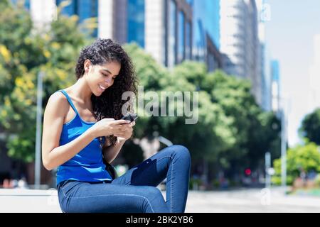 Giovane adulta brasiliana che invia messaggio con telefono in città in estate Foto Stock