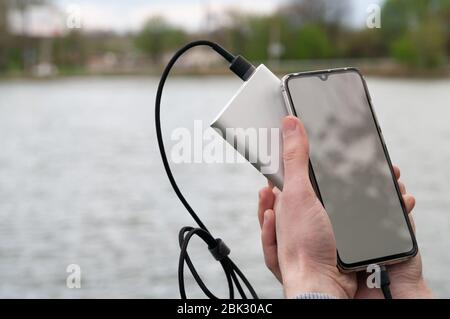 Man utilizza uno smartphone con schermo bianco vuoto durante la ricarica dalla banca di alimentazione sulla riva del fiume. Concetto tecnologico moderno. Messa a fuoco selettiva. Foto Stock