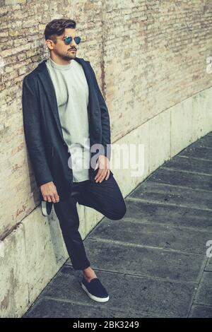 Bell'uomo elegante appoggiato su una parete con occhiali da sole e  acconciatura moderna e barba. Esterno in strada. T-shirt bianca e giacca  nera Foto stock - Alamy