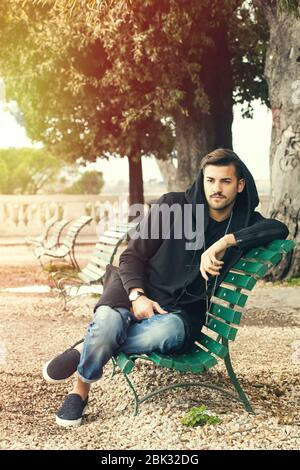 Giovane e alla moda, rilassante su una panchina in un parco con alberi. Il ragazzo si veste di moda, indossando parzialmente una felpa con cappuccio. All'aperto in un parco. Foto Stock