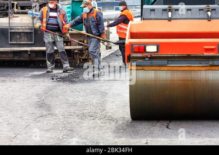 Il team di lavoro dei lavoratori della strada in maschere mediche protettive e tute depongono un nuovo rivestimento di asfalto sul cantiere. Foto Stock