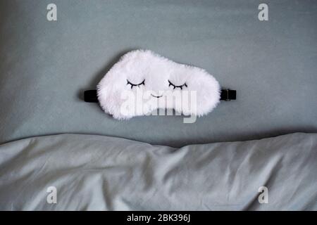 Maschera bianca cute sleep in soffice pelliccia sintetica a forma di nuvola con occhi chiusi ricamati su di essa su un cuscino e una coperta grigio blu. Foto Stock