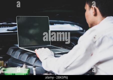 Il riparatore di auto indossa un'uniforme bianca che regge e tiene una chiave che è un attrezzo essenziale per un meccanico con il motore di controllo del laptop Foto Stock