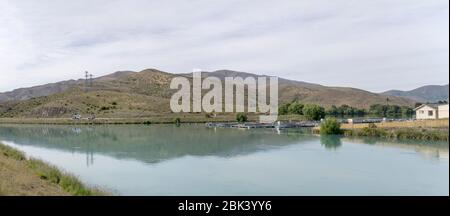 Paesaggio con allevamento di salmone galleggiante al braccio di Wairepo del lago Ruataniwha con piste sterili sullo sfondo, girato in una luce nuvolosa primavera brillante vicino a T. Foto Stock