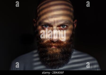 Ritratto ravvicinato di un uomo barbuto di mezza età con occhi espressivi e ombre a righe sul suo volto con sfondo nero Foto Stock