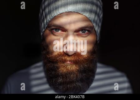 Ritratto ravvicinato di un uomo barbuto di mezza età con occhi espressivi e ombre a righe sul suo volto con sfondo nero Foto Stock