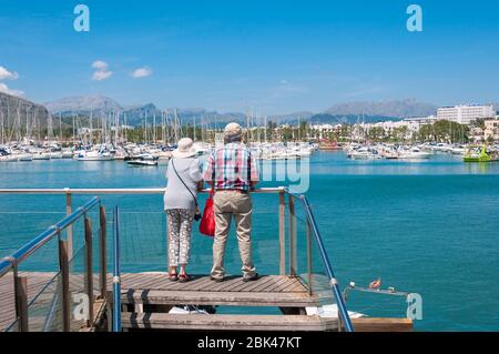Pollensa, Isole Baleari/Spagna; maggio/19/2018: Coppia di turisti sulle spalle, guardando il molo nel porto di Pollensa in una giornata di sole Foto Stock