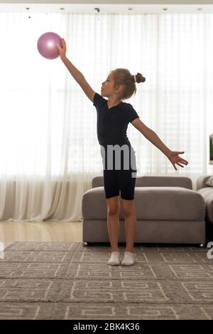 La ragazza con gli occhi blu Toddler fa ginnastica ritmica in un body nero, gioca con una palla rosa sulla casa Foto Stock