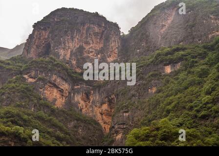 Wuchan, Cina - 7 maggio 2010: Dawu o Misty Gorge sul fiume Daning. Alte enormi scogliere nere e marroni con fogliame verde in macchie sotto il cielo d'argento. Foto Stock