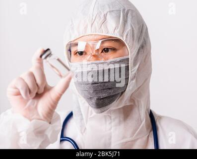 Donna asiatica medico o infermiere in uniforme PPE e guanti con maschera protettiva in laboratorio che tiene il flacone di vaccino Foto Stock